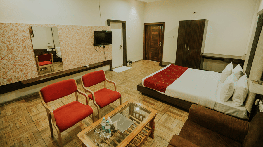 A bedroom at TGI Jahaj Mahal, Mandu contains a king size bed, 2 chairs, a glass table, and wall-mounted television.