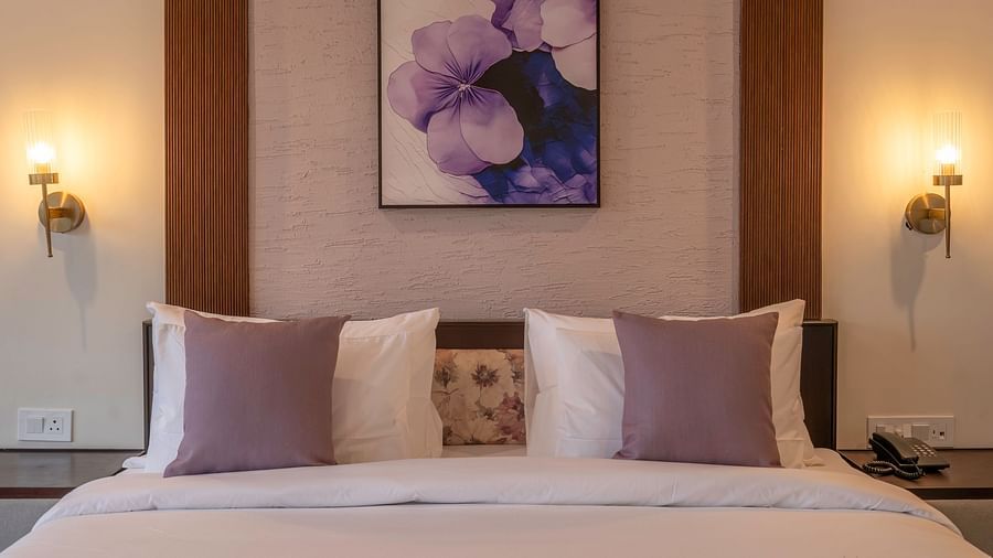 View from the foot of the bed with neatly made sheets and pillows, bedside lamps on both sides, and a floral painting above the headboard at The Chamomile Estate.