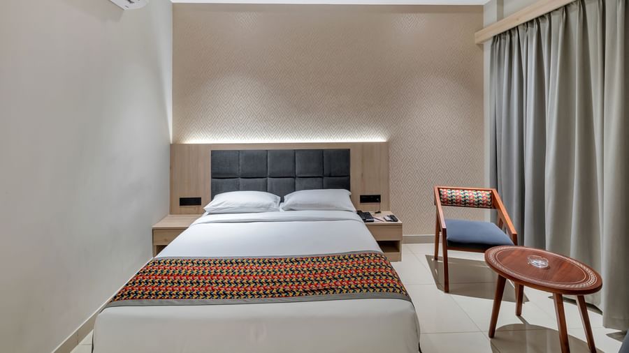 A cosy hotel room with a double bed featuring a colourful patterned throw, bedside tables, a chair, a small round table, and an air conditioning unit | The Citi Residenci Hotel Banskopa