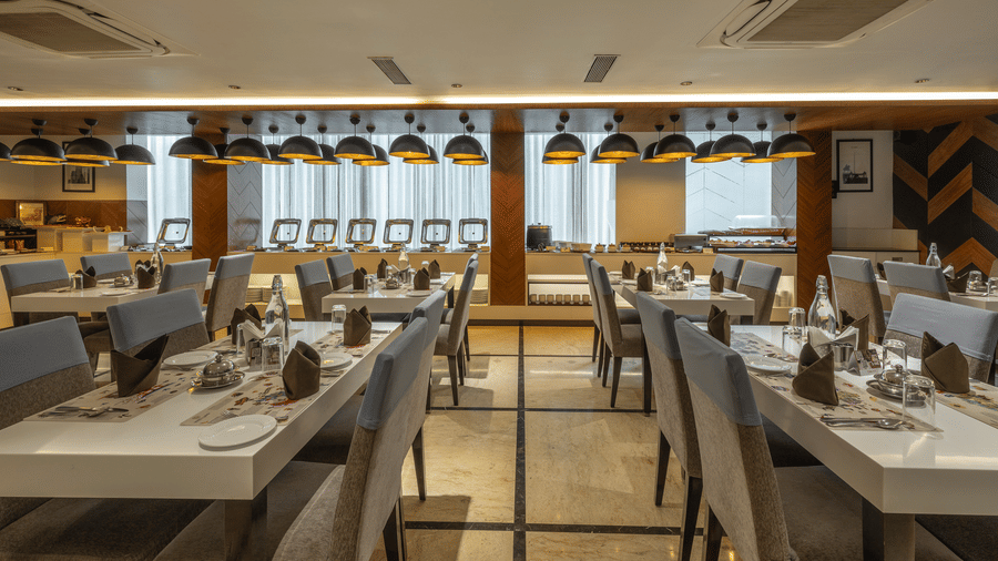 Restaurant dining hall at The Citrine, Bangalore, with rows of white tables set with napkins, grey chairs, and dome pendants over a buffet line.