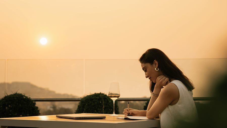 A woman seated at an outdoor table facing the horizon, with a glass placed on the table and the sun setting in the background at The Evren, Vagator.