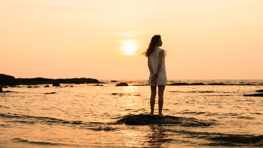 A woman standing in shallow seawater facing the horizon, with the sun low in the sky and waves surrounding her at The Evren, Vagator.