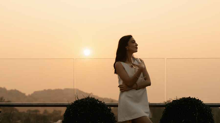 A woman standing near a glass railing facing the horizon, with the sun low in the sky and hills visible at The Evren, Vagator.