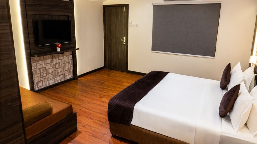 Hotel room interior featuring a bed with dark brown and white bedding, a flat-screen TV mounted on a wooden panel, wooden flooring, and an air conditioner above a covered window - The Grand Legacy Resort & Spa