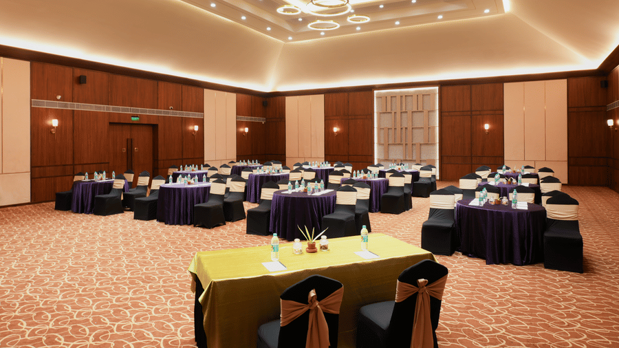 Banquet hall at Vedic Village Spa Resort Kolkata arranged with round tables in green and blue linen, black chair covers, and decorative coffered ceiling with warm lighting.