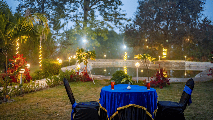 Outdoor dining setup at the lawn of Vedikant Resorts The Mallard Corbett featuring a table for two by the pool side and the entire space is decorated with fabric pieces and lights wrapped on tree trunks and branches.
