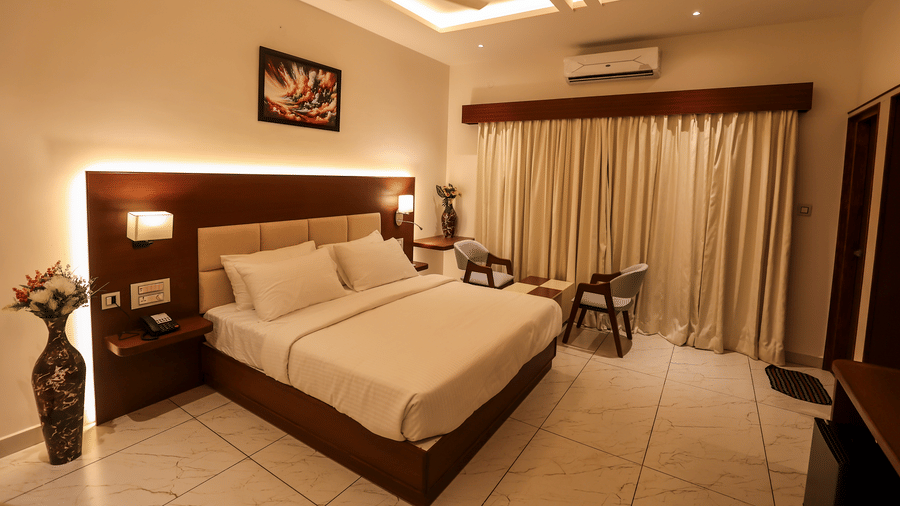 A hotel bedroom featuring a double bed, an illuminated wooden headboard, a wall picture frame, a flower vase, and heavy window curtains | Lords Sahyadri Resort, Shivamogga