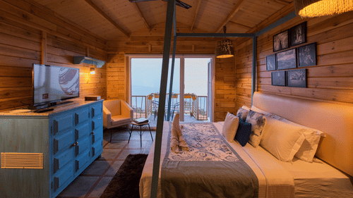 A bedroom at Polo Cherrapunjee Resort with a bed, a colourful dresser, seating area, and glass doors opening to a balcony with scenic views.