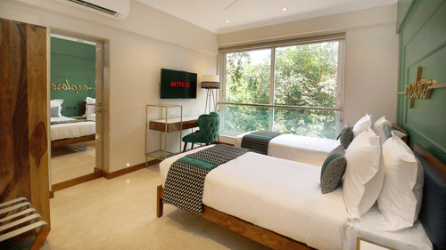 A twin bed with a view outside, accompanied by a wardrobe and mirror on the side, a small table, a chair, and a TV on top