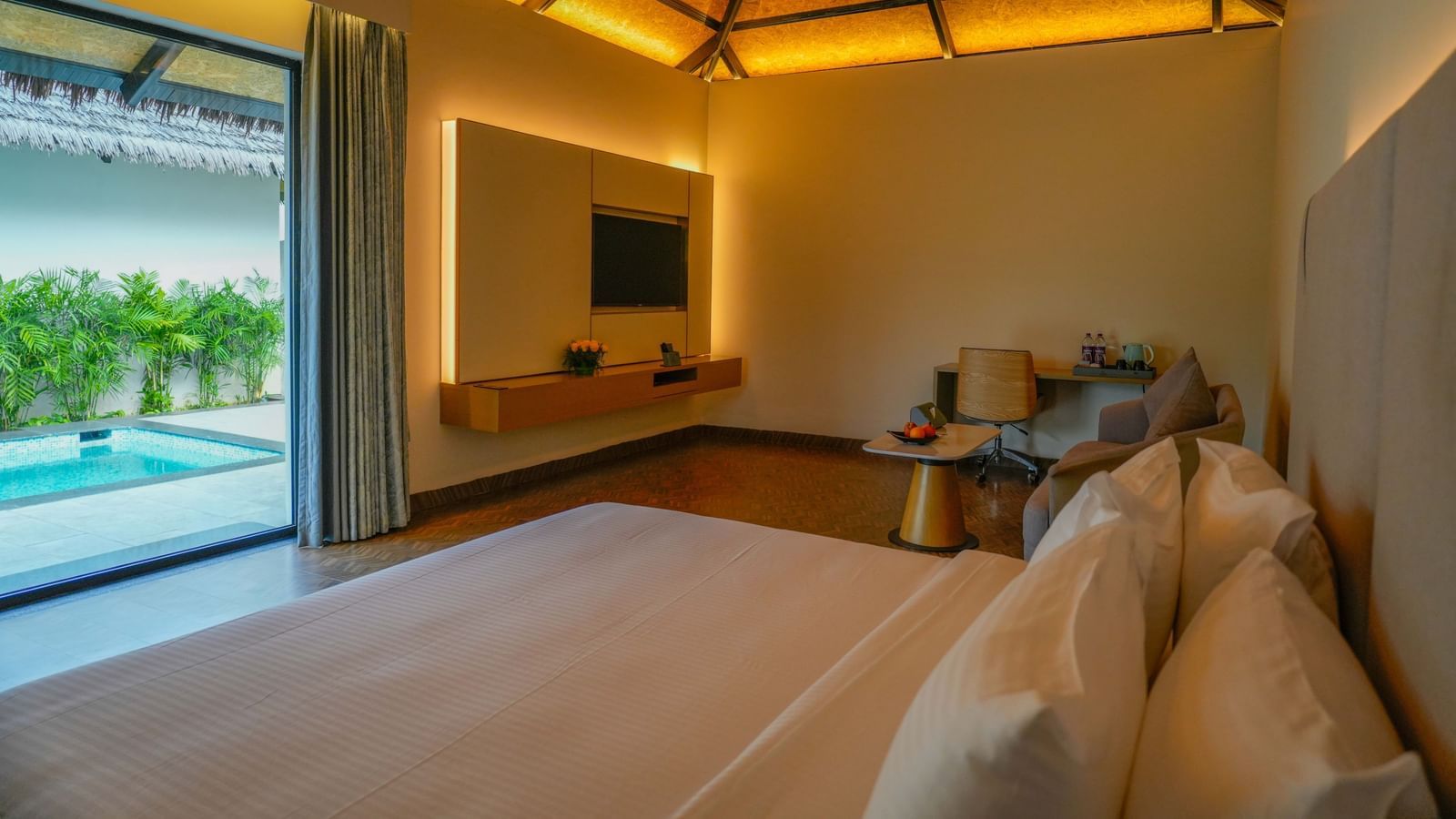 Interior view of a luxury villa room with a neatly made bed, warm ambient lighting, a mounted TV, a sitting area with a desk and chair, and a view of a private pool through the open sliding glass door - The Shore Trishvam, Pondicherry