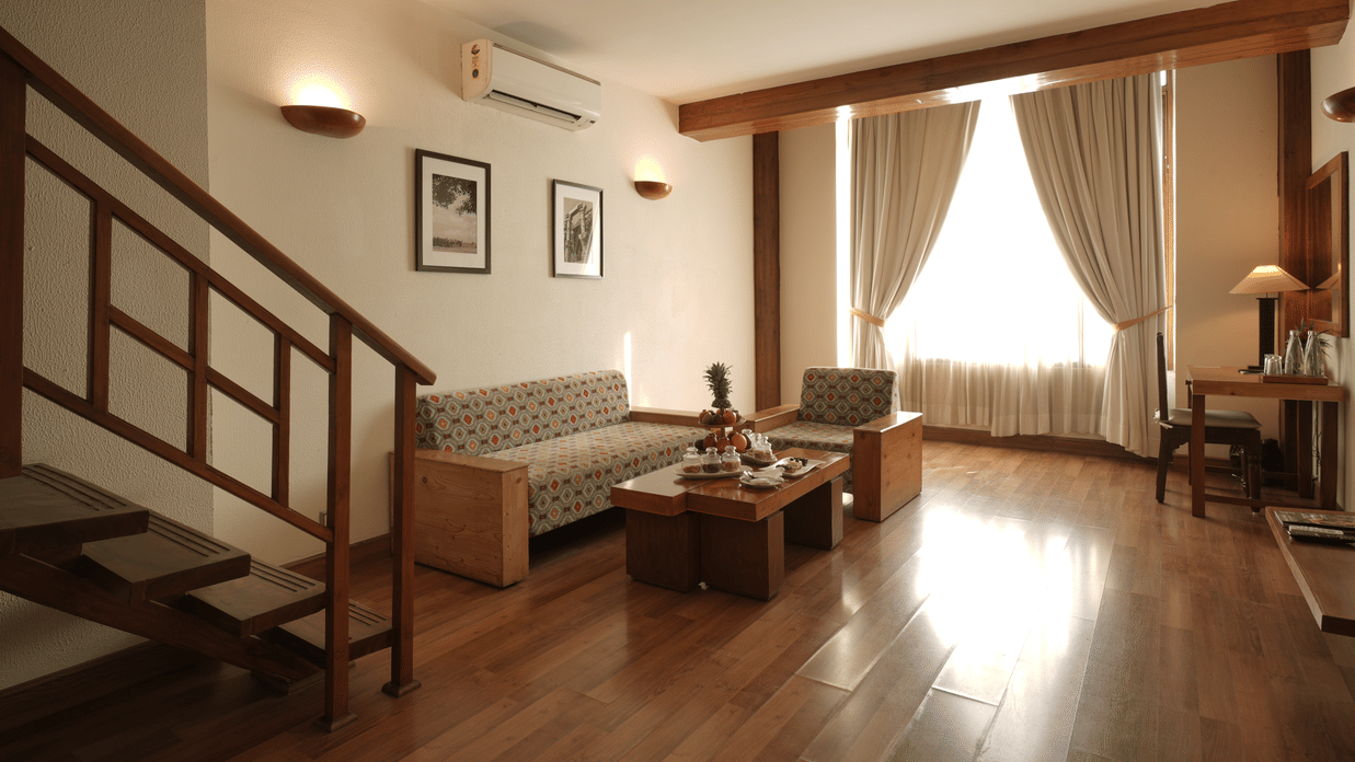 A duplex-style living area with a staircase, sofa seating, wooden flooring, and large windows allowing natural light in the Penthouse Suite at Clarks Amer, Jaipur.