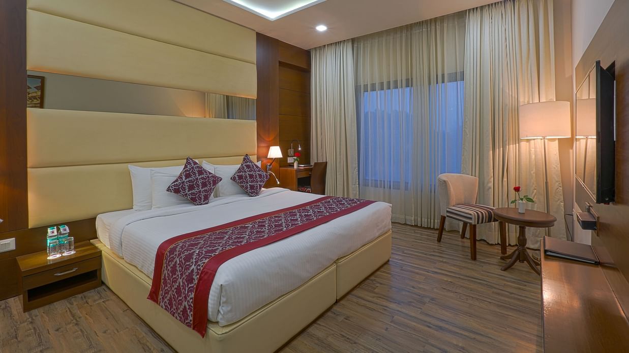 Room at Clarks Inn Suites, Near Gwalior DD Mall with double bed, side table, desk, chair, TV, window with curtains, armchair, round table, and wooden flooring.