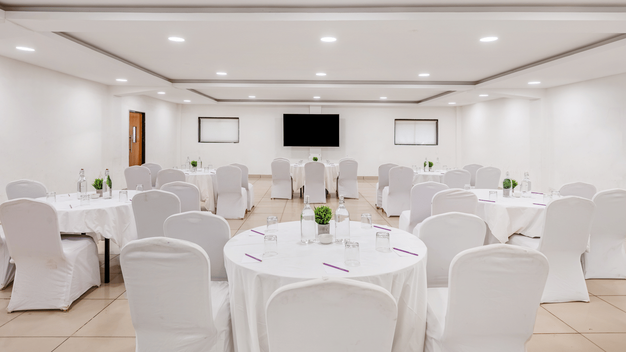 Bright conference room at Nature Trails Durshet with round tables, white chair covers, a wall-mounted TV, and recessed lighting.