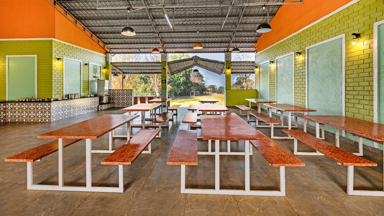 Nature Trails Kudalika dining hall with red-topped bench tables, tiled buffet counter on the left, green brick walls, coloured pendant lights and open garden view.