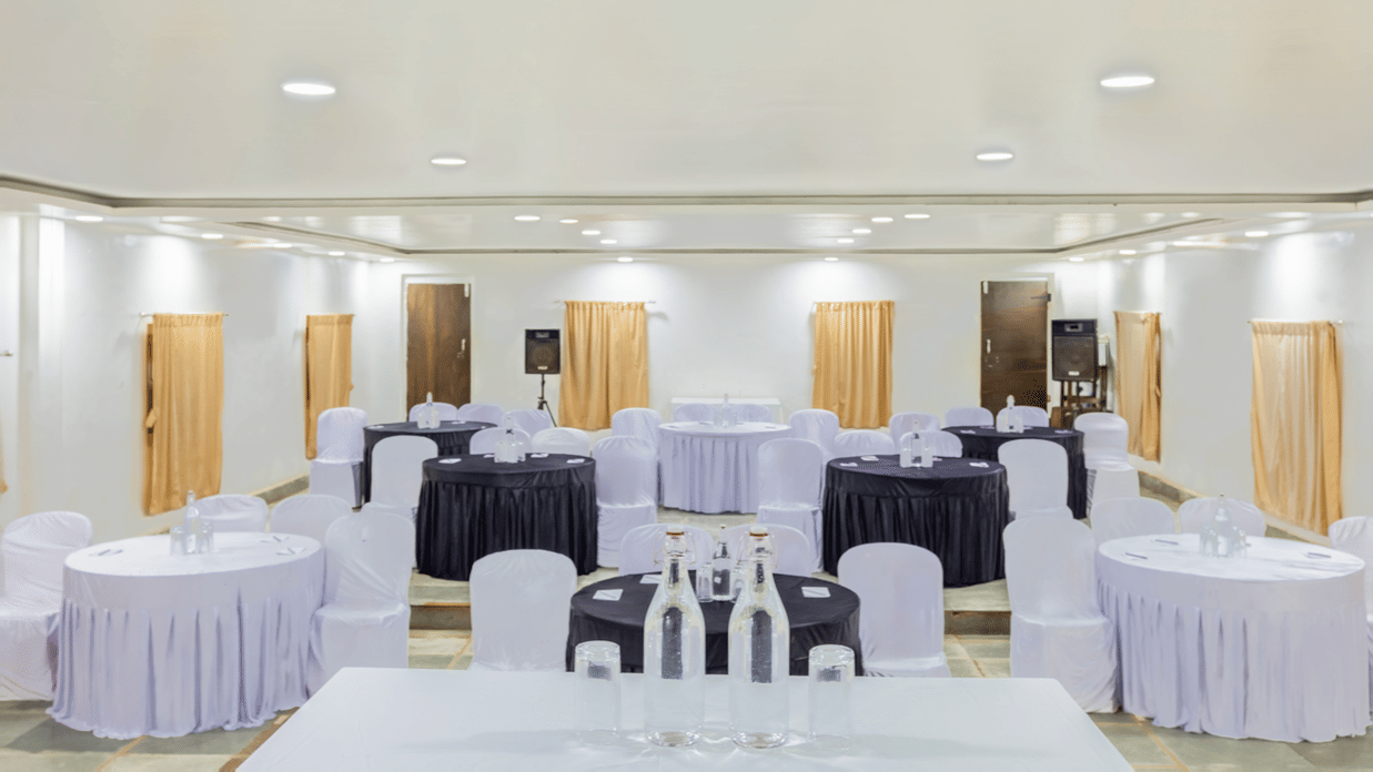 Spacious banquet hall with round tables in white and purple draping, white chair covers, warm recessed lighting and wooden partition walls in background.