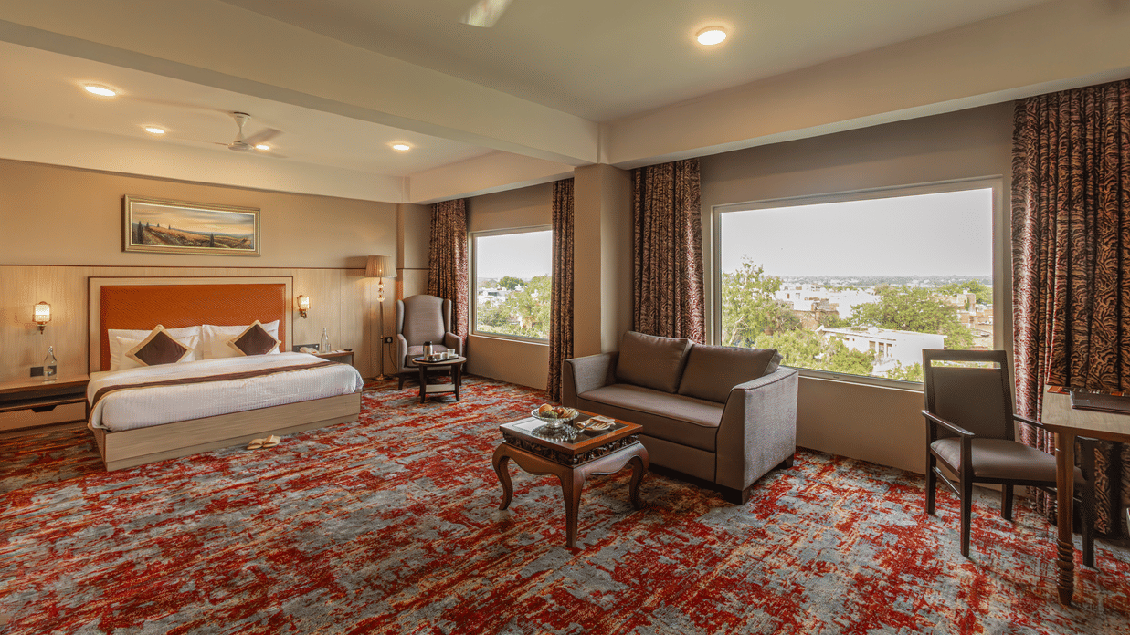 The Junior Suite Room at The Lalita Grand with a double bed, ceiling lights, a 2-seater sofa, ceiling lights, and a window overlooking the greenery.