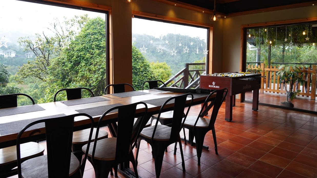 A spacious indoor dining or common area with long wooden tables and black chairs, featuring large windows that offer expansive views of the misty, green mountainous landscape outside. The floor is tiled, and the ceiling has wooden beams. | The Wild Trails 