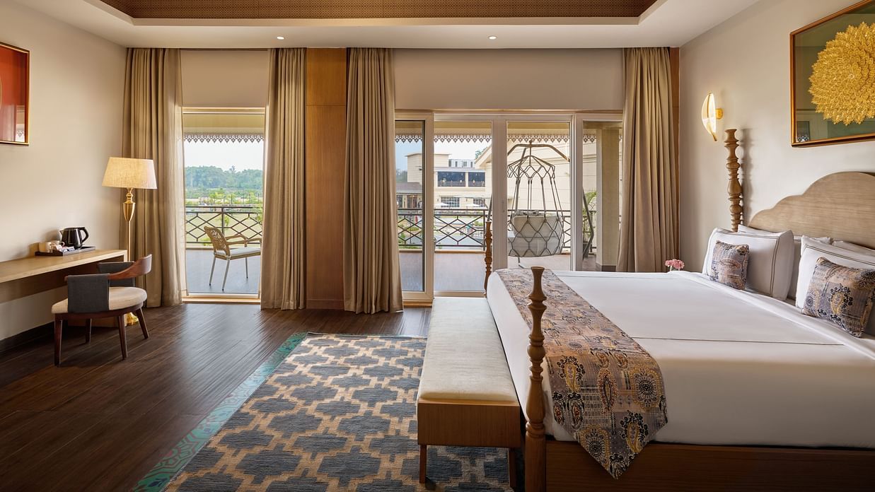A well-lit hotel room featuring a king size bed with a patterned throw, a wooden desk and chair, and a balcony with a swing offering a view of a cityscape. The room has a wooden ceiling and a patterned rug - Voyage The Solitaire Bliss.