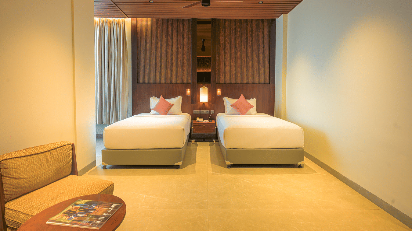 Twin bedroom with two neatly made beds, red accent cushions, wooden ceiling, bedside furnishings and a patterned rug.