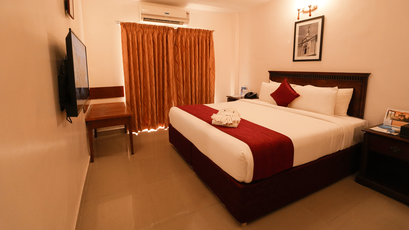 MGM Vailankanni Residency - Deluxe Room - Queen Bed. Image of the well-made bed, bedside tables, and floor-to-ceiling curtains.