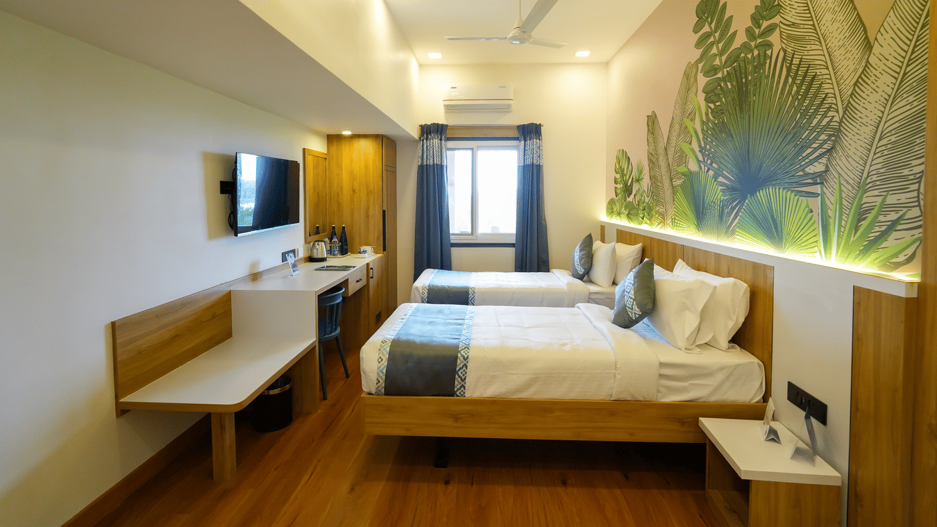 A contemporary room with two beds, light wood furniture, and a colorful mural of tropical leaves behind the beds.