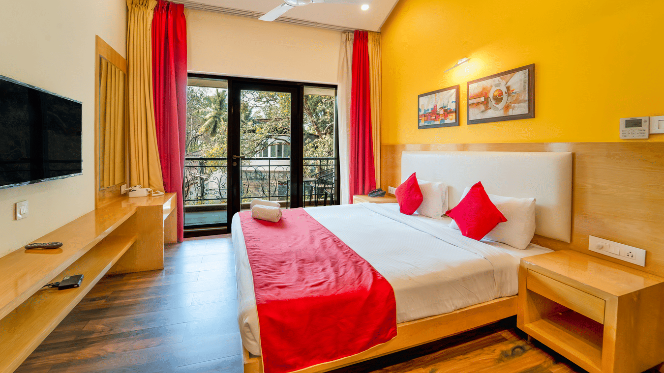 Rosetum Anjuna Deluxe Room with a king bed, red runner, red accent pillows, yellow walls, framed artwork, flat-screen TV, and open balcony doors with garden views.