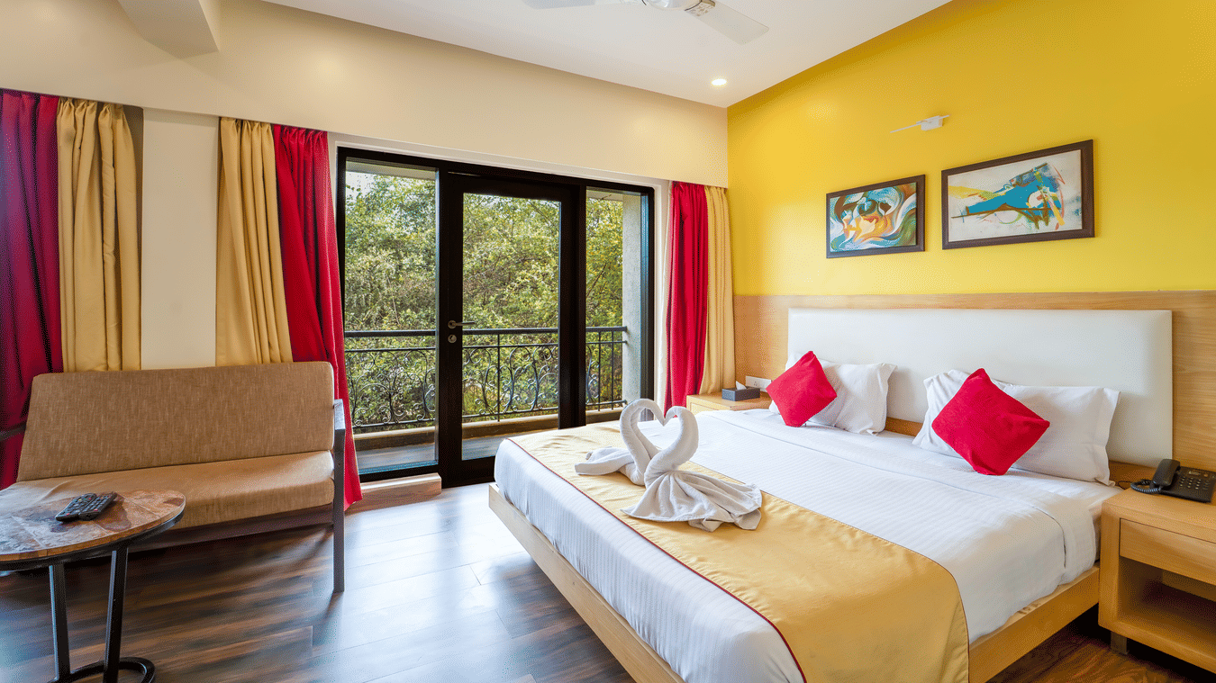 Rosetum Anjuna's Executive Suite bedroom featuring a king bed with towel swans, red cushions, balcony with garden view, yellow accent wall, and framed artwork above the headboard.