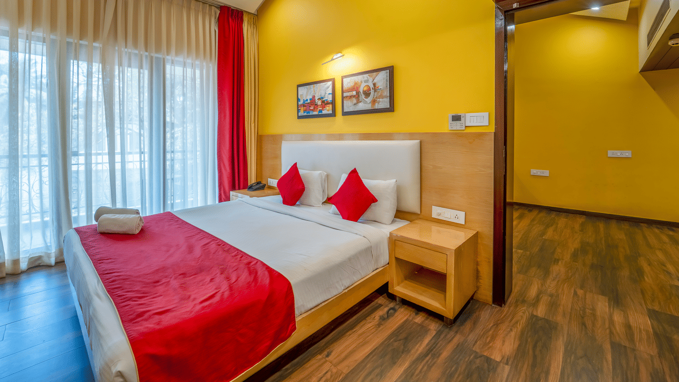 Rosetum Anjuna Royal Suite bedroom with a king bed, red accent pillows, yellow walls, framed artwork, sheer curtains, balcony access, and wooden flooring.