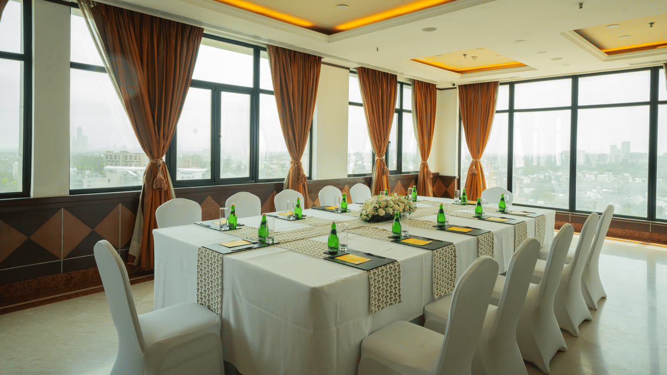 Sampan Tea Room at The Suryaa set with rectangular tables, white covers, green bottles, notepads, pens, warm recessed lighting, large windows with city view and brown curtains.