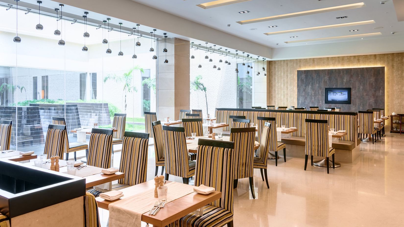 A spacious and contemporary dining area with striped seating, large windows offering a view, and soft pendant lighting - Balaji Sarovar Premiere, Solapur