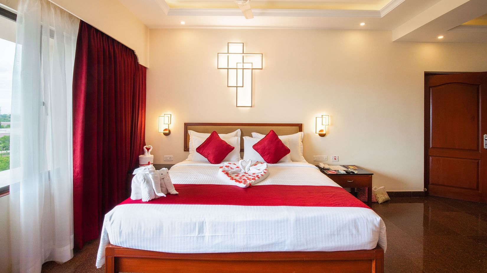 A neatly made bed in Honeymoon Suite with bedding, two pillows, and bedside tables, in a well-lit room with large windows and curtains