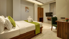 A hotel room featuring a neatly made bed with white linens and green accents, a towel arrangement, wooden wardrobe, desk with chair, wall-mounted TV, and soft ambient lighting - Magnus Fortune House, Magarpatta