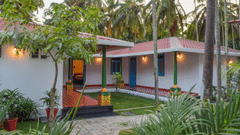 A white resort cottage with a red roof and blue window frames, surrounded by palm trees and green lawns - Ibex Resorts, Coimbatore (Kakarla)