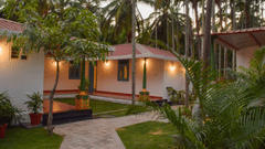 A white resort cottage with a red roof and blue window frames, surrounded by palm trees and green lawns - Ibex Resorts, Coimbatore (Kakarla)
