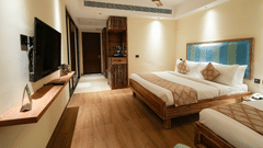 View of the room showcasing the television on a wooden shelf, a mini fridge cabinet, and a glimpse of the entry door, with wooden flooring and beige walls.