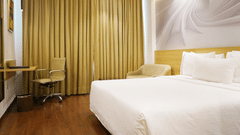 A corner view of a spacious guest room at Nemesia featuring a comfortable bed, a work desk, a seating area, and floor-to-ceiling curtains, creating a relaxing and inviting atmosphere.