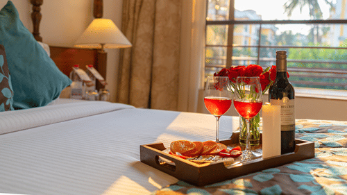 A close-up image of a bed featuring a tray that holds two half-filled wine glasses, a wine bottle, a candle, roses, and snacks.