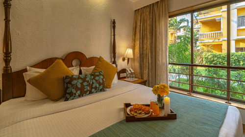 An image of a room showcasing a beautifully decorated bed adorned with a tray of candles, flowers, and fruits, complemented by a large window.