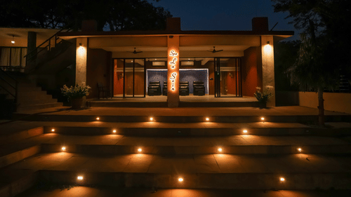 Exterior night view of the main building at Adamo The Village showing a modern structure with floor-to-ceiling glass doors and series of steps leading up to the entrance lined on both sides by small ground lights.