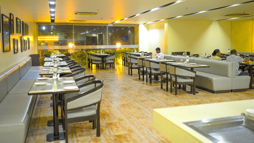 A restaurant dining room at Ambar Sarovar Portico, Gandhidham features neatly arranged tables, chairs, and cushioned booth seating along the wall.