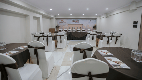 A well-lit banquet hall with elegant seating, white chair covers, and neatly arranged dining tables - Biznotel By Pride Surat