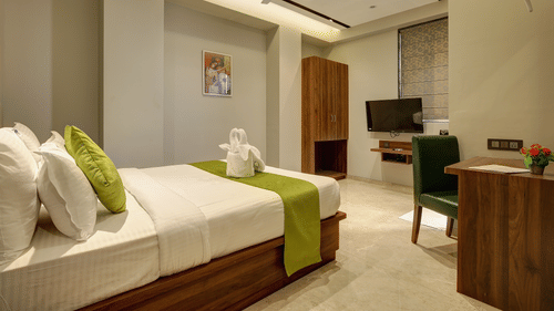 A hotel room featuring a neatly made bed with white linens and green accents, a towel arrangement, wooden wardrobe, desk with chair, wall-mounted TV, and soft ambient lighting - Magnus Fortune House, Magarpatta