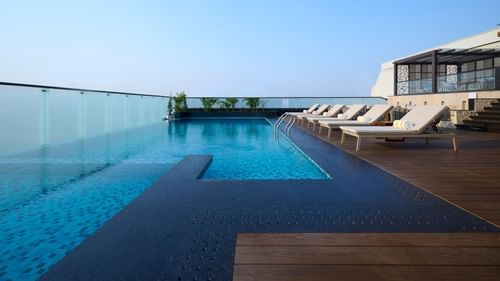 A rooftop infinity pool with clear blue water extends to the edge of the building, offering a view of the clear sky, with lounge chairs lined up on the decking. - Iris Sarovar Portico, Sector 85, Gurugram