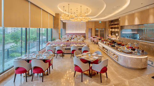 A restaurant interior at KPG Sarovar Portico, Raj Nagar Extension, Ghaziabad featuring dining tables, padded chairs, a long buffet counter, and window blinds.