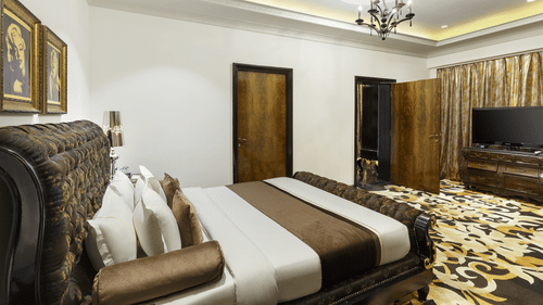 The spacious presidential suite bedroom with a king size bed, upholstered headboard, bedside tables, chandelier lighting, television unit and patterned flooring at La Marvella, Bengaluru.