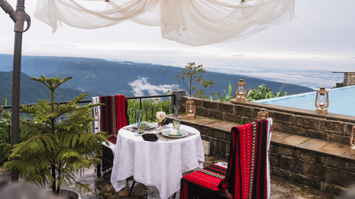 A dining table with two chairs on either side with a view of waterfalls in broad daylight - Polo Cherrapunjee Resort