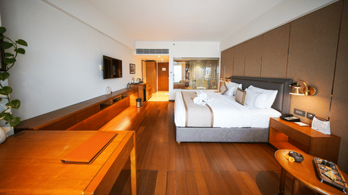 A perspective view inside the Premium King Room at Pride Premier Udaipur. The space displays a desk in the foreground, timber flooring, a double bed with pillows against the wall, a television mounted on the wall, and a doorway in the distance.