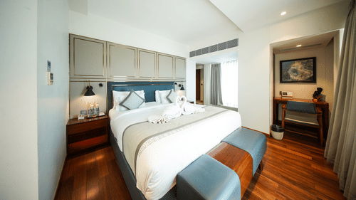 An angled view of the bedroom area in the Premium Suite Room at Pride Premier Udaipur. The space displays a double bed with linen, bedside tables, lamps, and an opening leading towards a desk and chair.