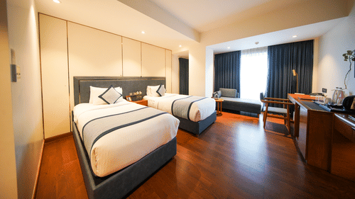 An angled view across the Premium Twin Room at Pride Premier Udaipur. The space displays two single beds arranged in sequence along the wall. A desk with a chair and a wall fixture are positioned on the opposite side near the window area.
