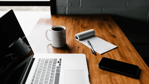 a laptop kept on a table with a mobile phone, notepad, a pen and a coffee mug